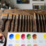 A set of professional paint brushes arranged by size and shape on a wooden work surface with paint swatches